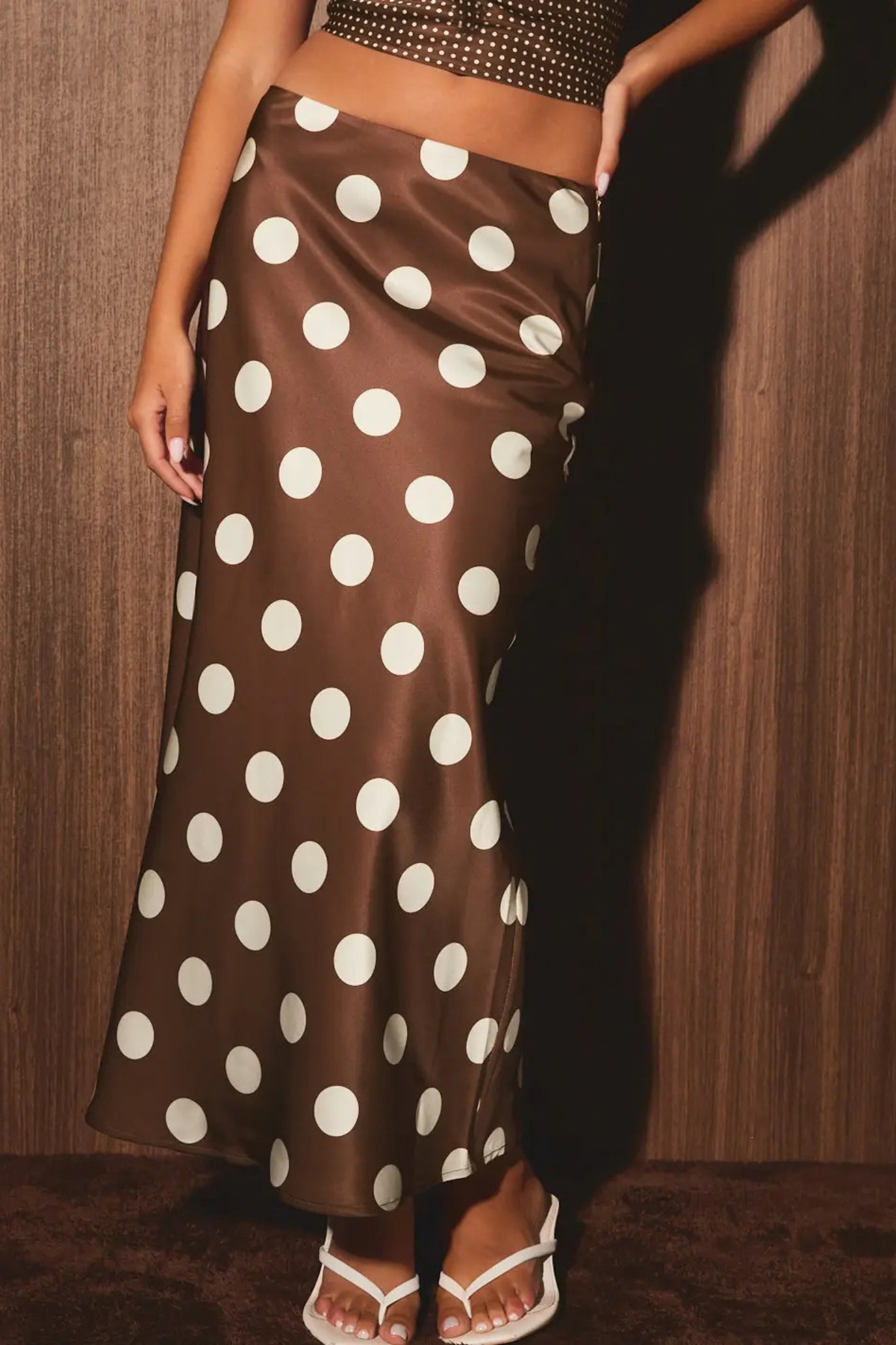 A brown satin maxi skirt with large white polka dots, worn by a model standing against a dark wood-paneled wall.