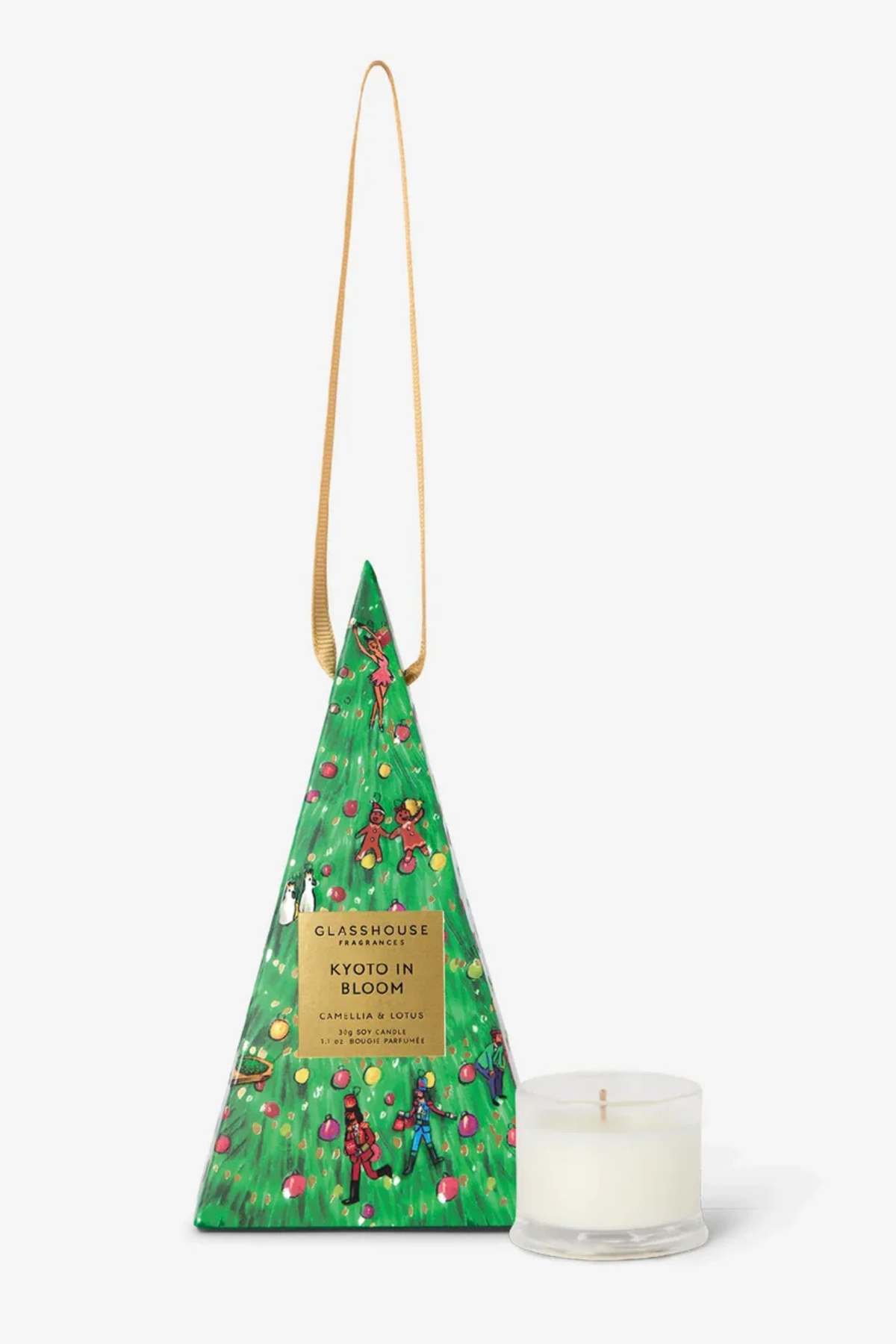 A triangular green gift box with a long tan strap, decorated with a Christmas tree pattern and a gold label reading 'Knots in Bloom', shown next to a small white candle in a clear glass jar on a light gray background.