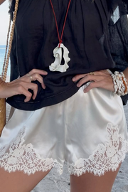 A close-up of a person wearing white satin shorts with a white lace hem, paired with a black top and a white shell pendant necklace.