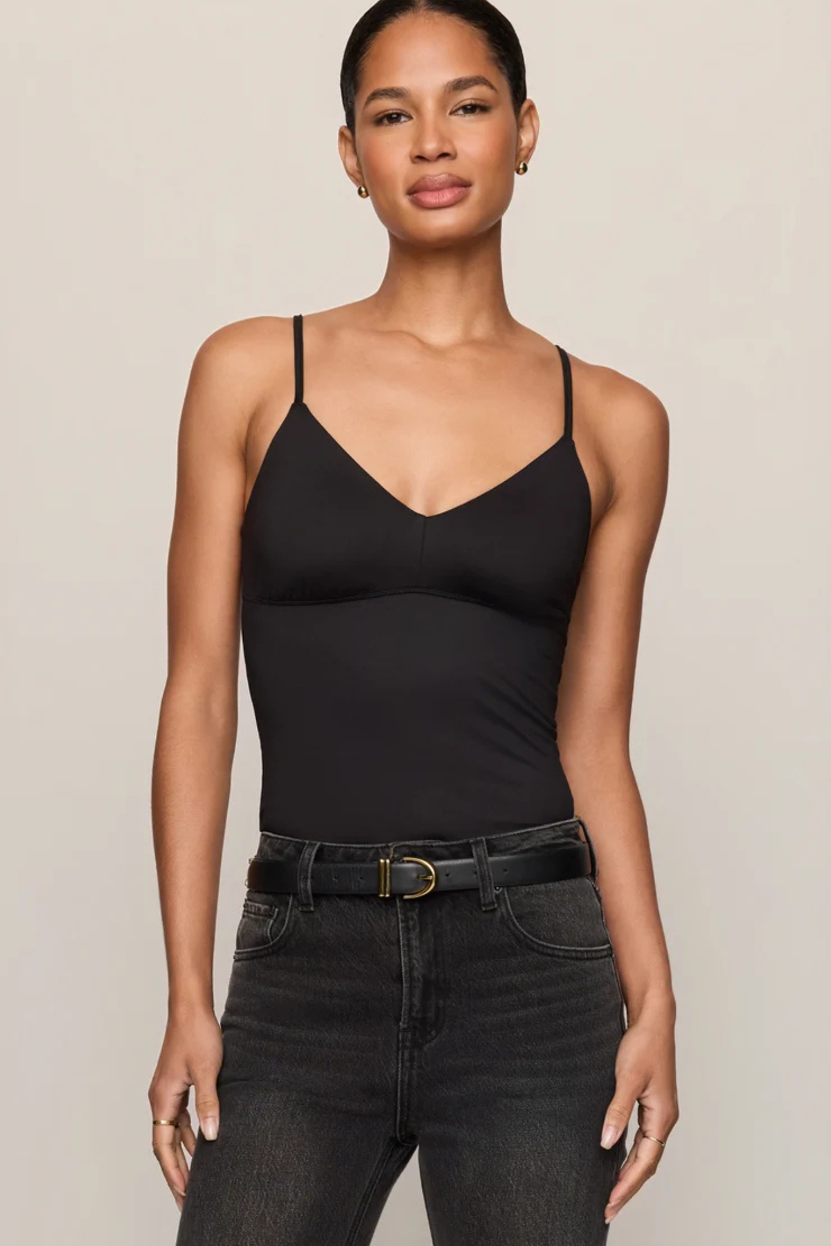 A woman modeling a black spaghetti-strap camisole top with a V-neckline, paired with dark gray jeans and a black belt, against a solid light beige background.