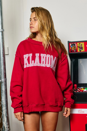 A woman wearing an oversized red sweatshirt with 'OKLAHOMA' printed in white arched letters across the chest, standing indoors next to a red arcade machine.