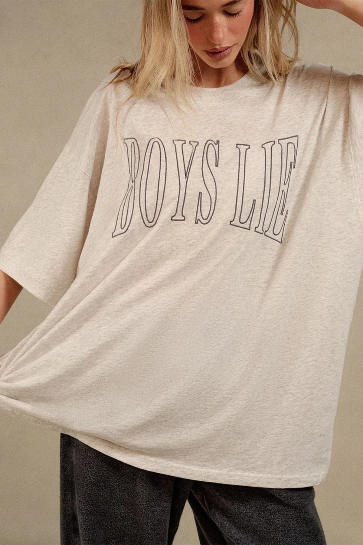 A woman wearing an oversized cream-colored t-shirt with 'BOYS LIE' printed in black outline across the chest, paired with dark gray jeans, against a neutral beige background.