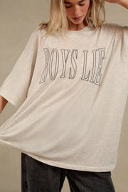 A woman wearing an oversized cream-colored t-shirt with 'BOYS LIE' printed in black outline across the chest, paired with dark gray jeans, against a neutral beige background.