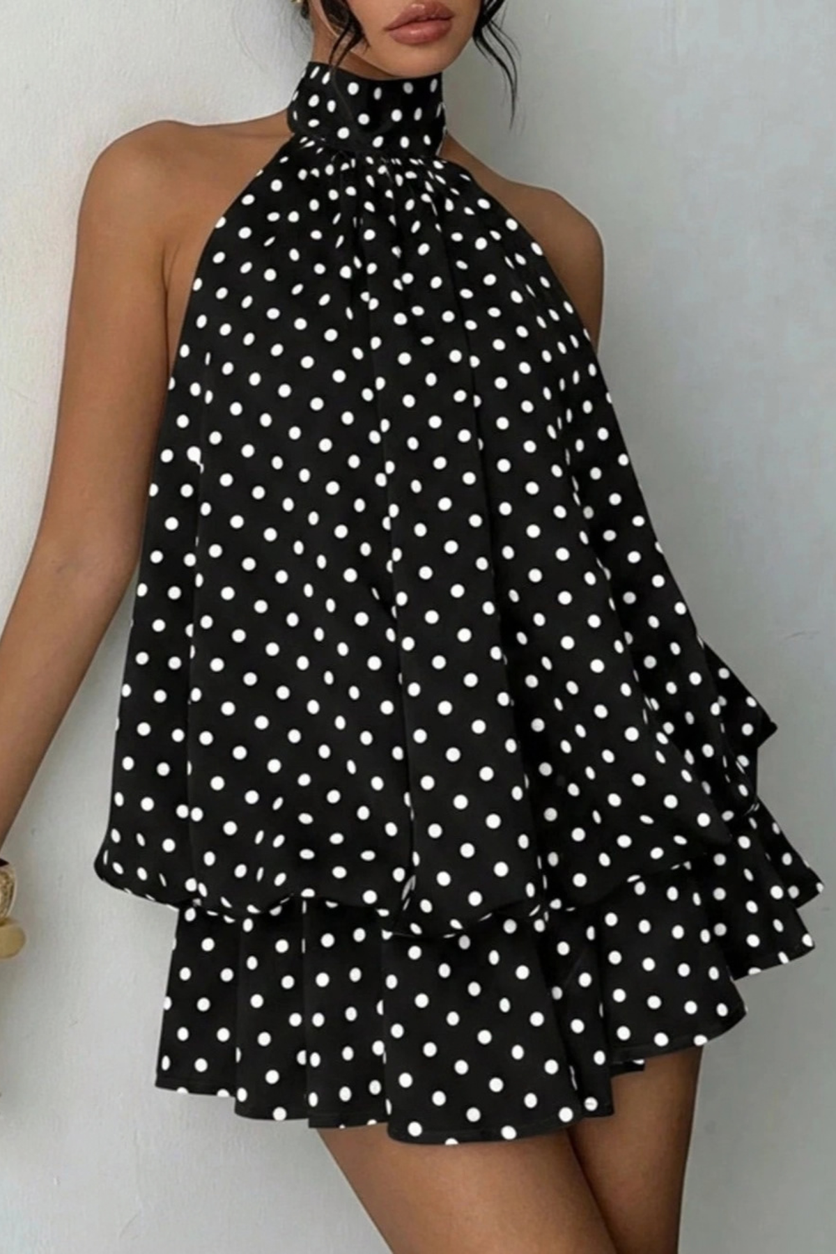 A sleeveless halter-neck dress with a black base and white polka dot pattern, featuring a high gathered neckline and layered ruffle hem, shown on a model against a plain light wall.