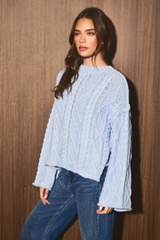 A woman wearing a light blue cable-knit sweater and blue jeans, posing against a brown wood-paneled wall.