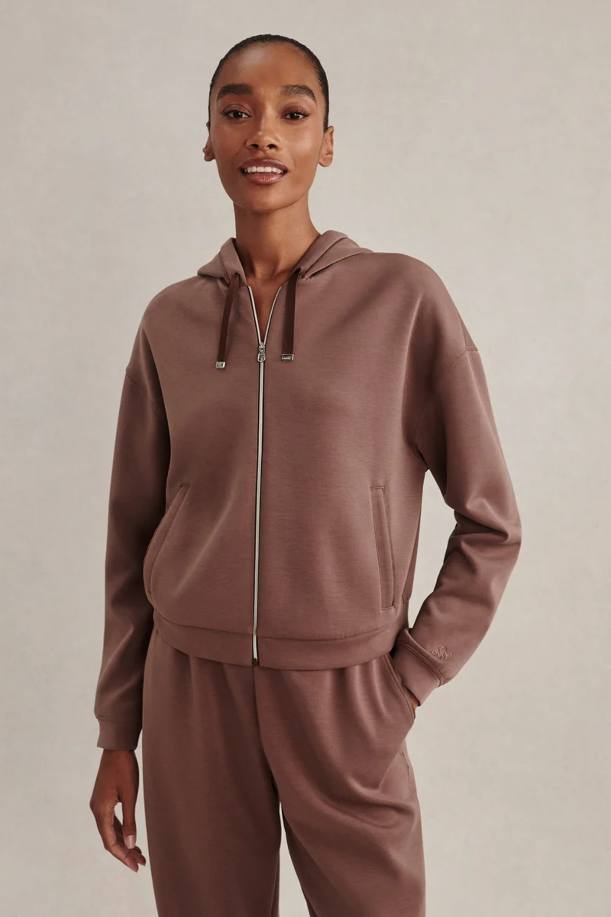 A model wearing a matching brown zip-up hoodie and sweatpants set against a plain beige studio background.