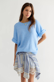 A woman modeling a light blue oversized knit sweater with rolled sleeves, paired with a blue and white striped shirt tied around her waist over white lace shorts, against a plain light gray studio background.