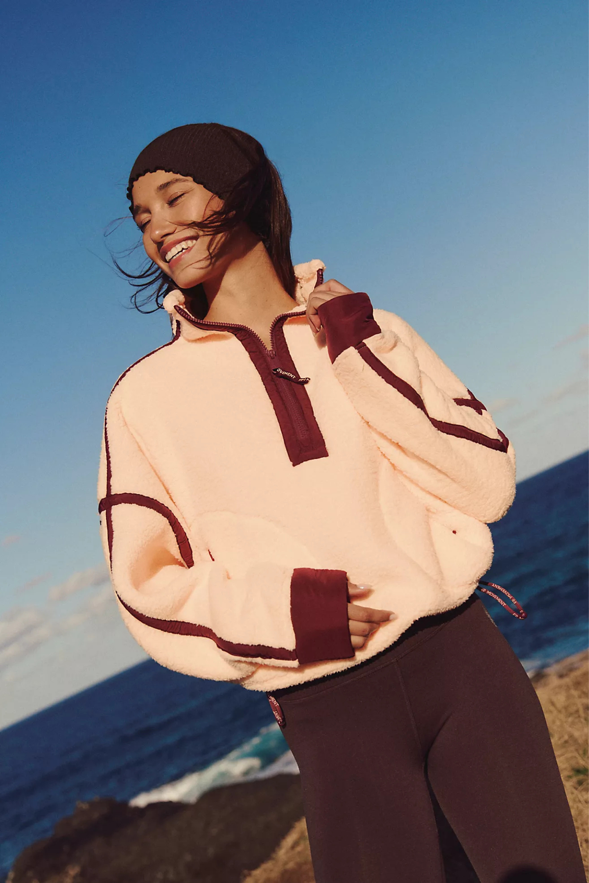A woman outdoors by the ocean wearing a cream fleece pullover with dark burgundy trim and matching dark leggings, smiling against a blue sky and sea background.