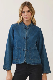 A woman modeling a cropped blue denim jacket with a mandarin collar, button-tab closure, and front patch pockets, paired with black pants against a plain light background.
