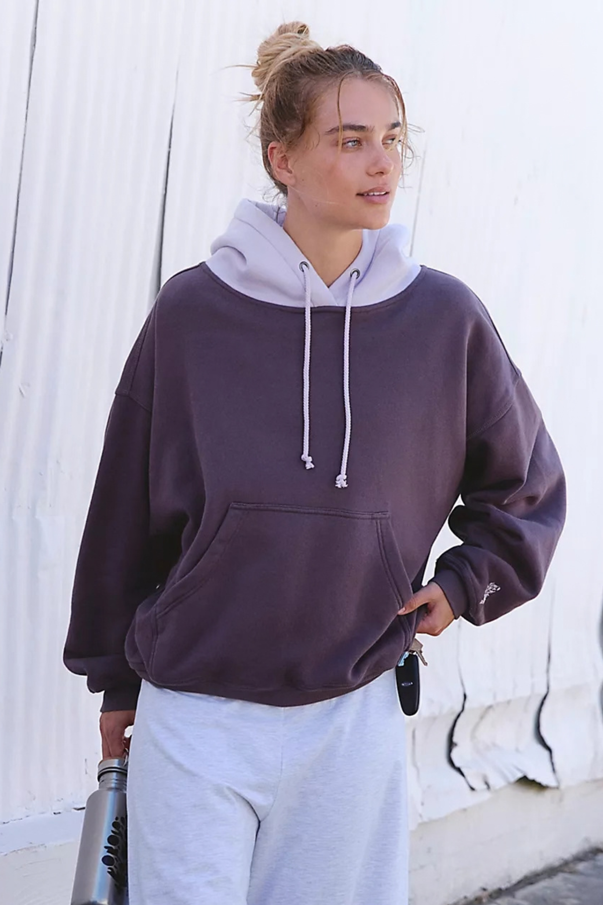 A woman wearing an oversized dark plum-purple pullover hoodie with a contrasting light lavender hood and white drawstrings, paired with light gray sweatpants, standing against a white corrugated metal wall.