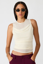 A model wearing a cream-colored sleeveless top with a sheer mesh yoke and ruched texture, paired with dark purple trousers and gold-framed sunglasses, against a light gray background.