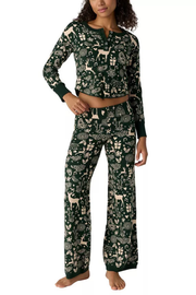 A woman modeling a matching two-piece pajama set with a dark green and cream holiday-themed print featuring deer, pine trees, and gift boxes, on a solid white background.