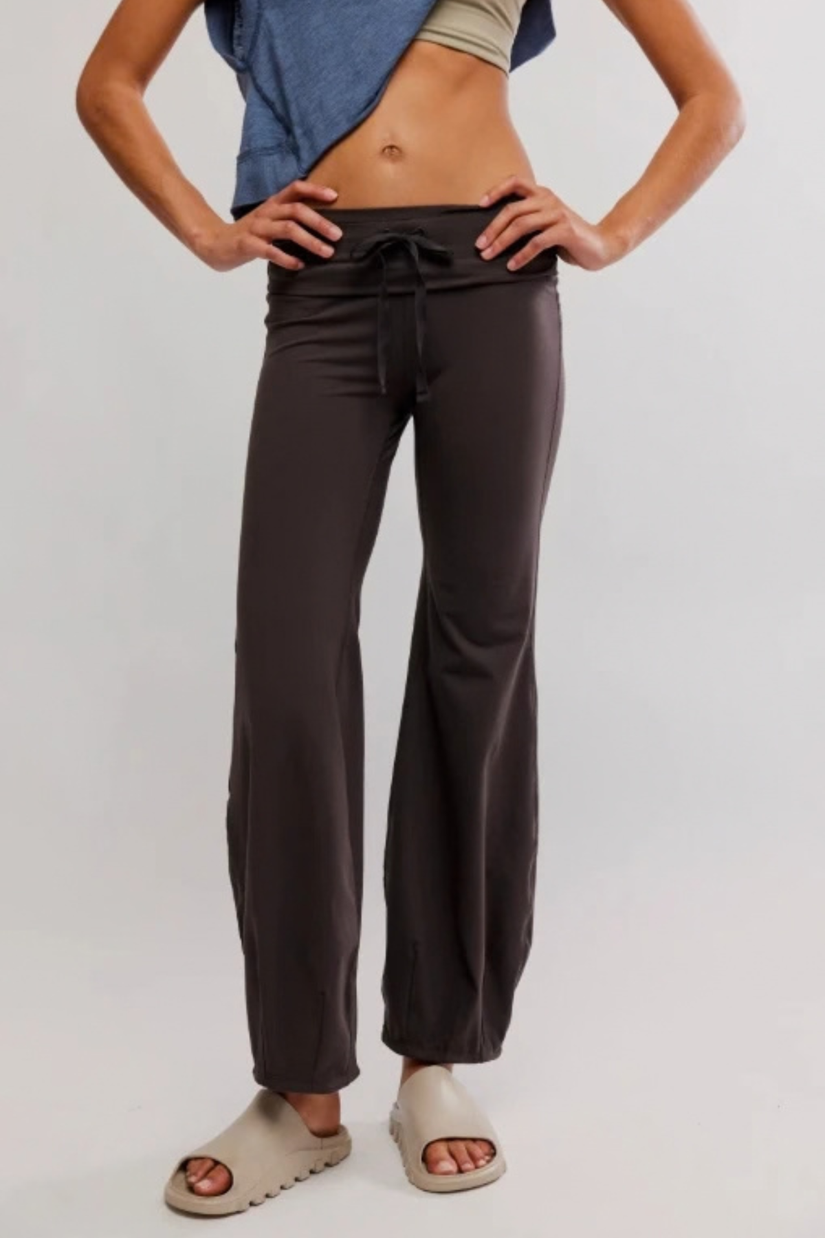 A model wearing dark brown wide-leg lounge pants with a drawstring waist, styled with a blue t-shirt and beige slide sandals against a light gray studio background.