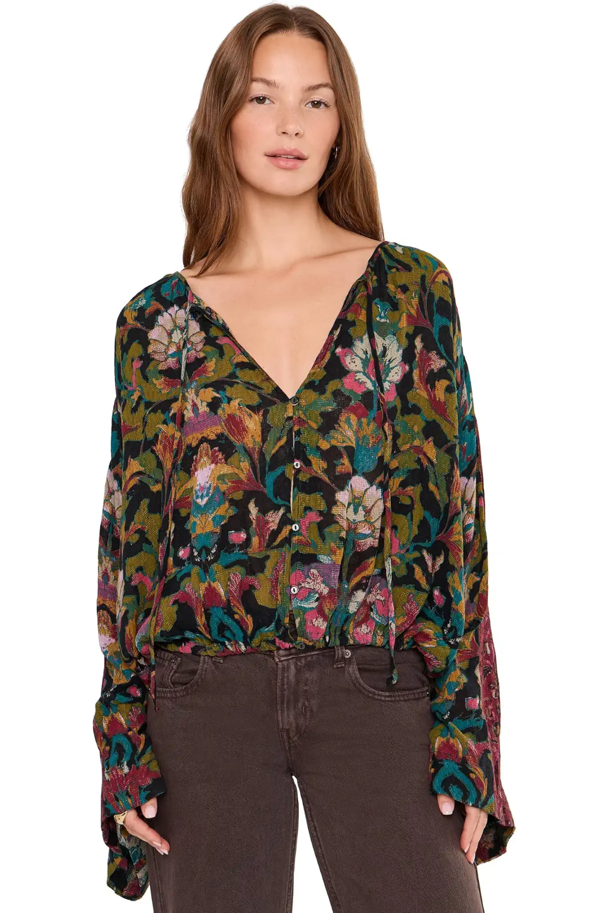 A woman modeling a long-sleeve button-front blouse with a dense floral and botanical print in olive green, burgundy, teal, and pink, paired with dark brown jeans against a plain white background.