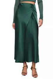 A dark green satin wrap skirt with a side tie, worn by a model with a matching green long-sleeve crop top and black strappy heels, against a light gray background.