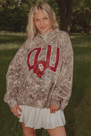 A woman standing outdoors in a grassy park wearing a brown and beige camouflage hoodie with a red and white Oklahoma 'OU' football helmet graphic on the front, paired with a white pleated skirt.