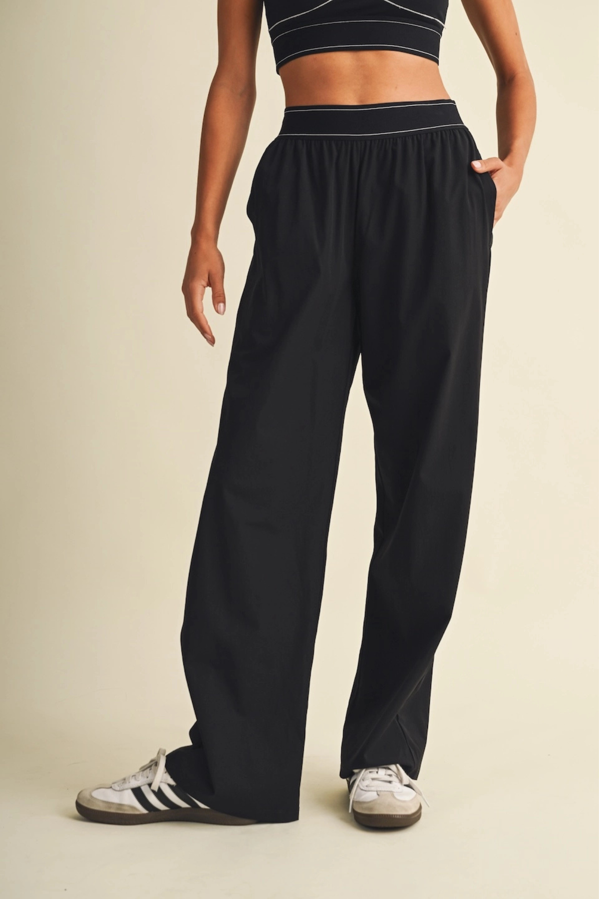 A person wearing loose black wide-leg sweatpants with a matching black crop top and white sneakers, standing against a plain beige studio background.