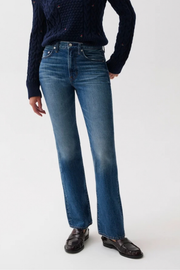 A model wearing a pair of medium-wash blue jeans with a subtle fade on the thighs, styled with a dark navy cable-knit sweater and dark brown leather loafers against a plain white studio background.