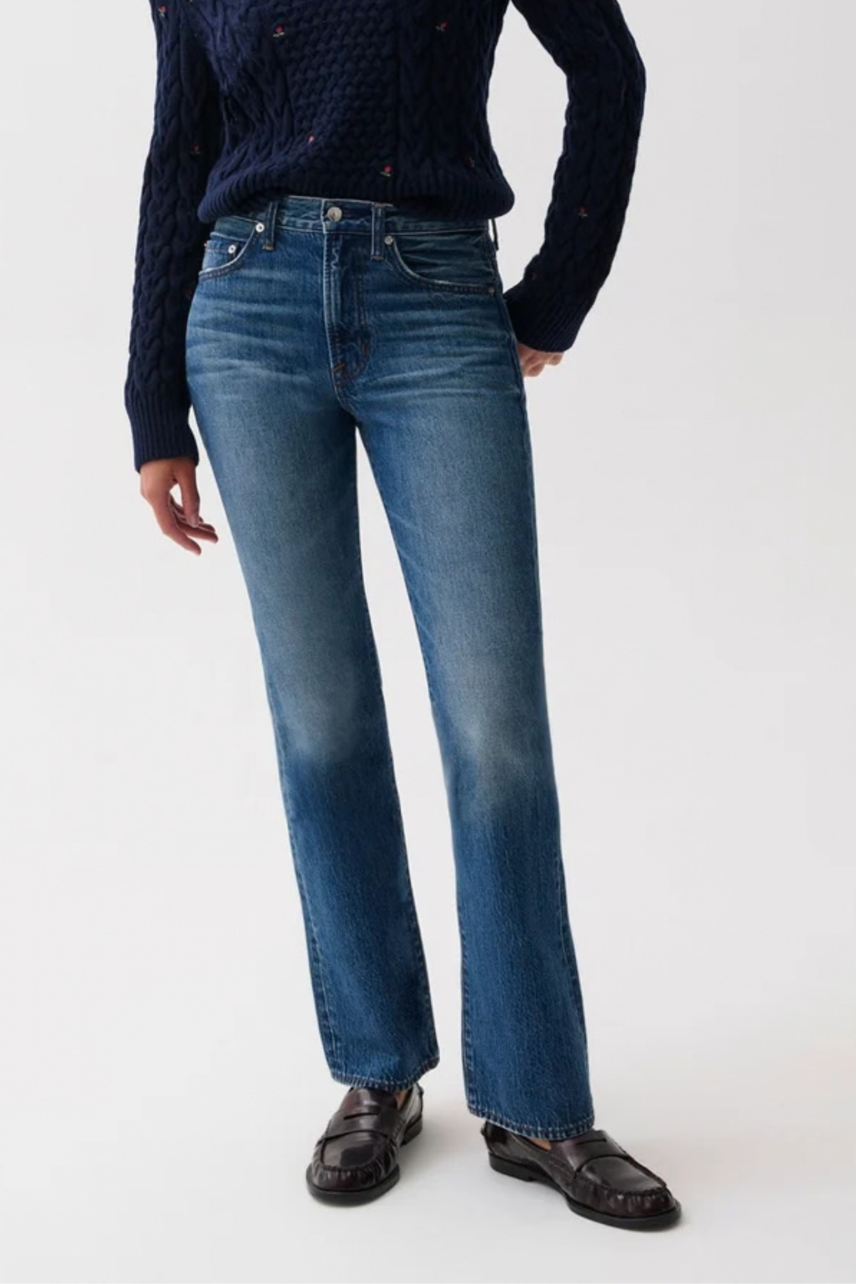 A model wearing a pair of medium-wash blue jeans with a subtle fade on the thighs, styled with a dark navy cable-knit sweater and dark brown leather loafers against a plain white studio background.