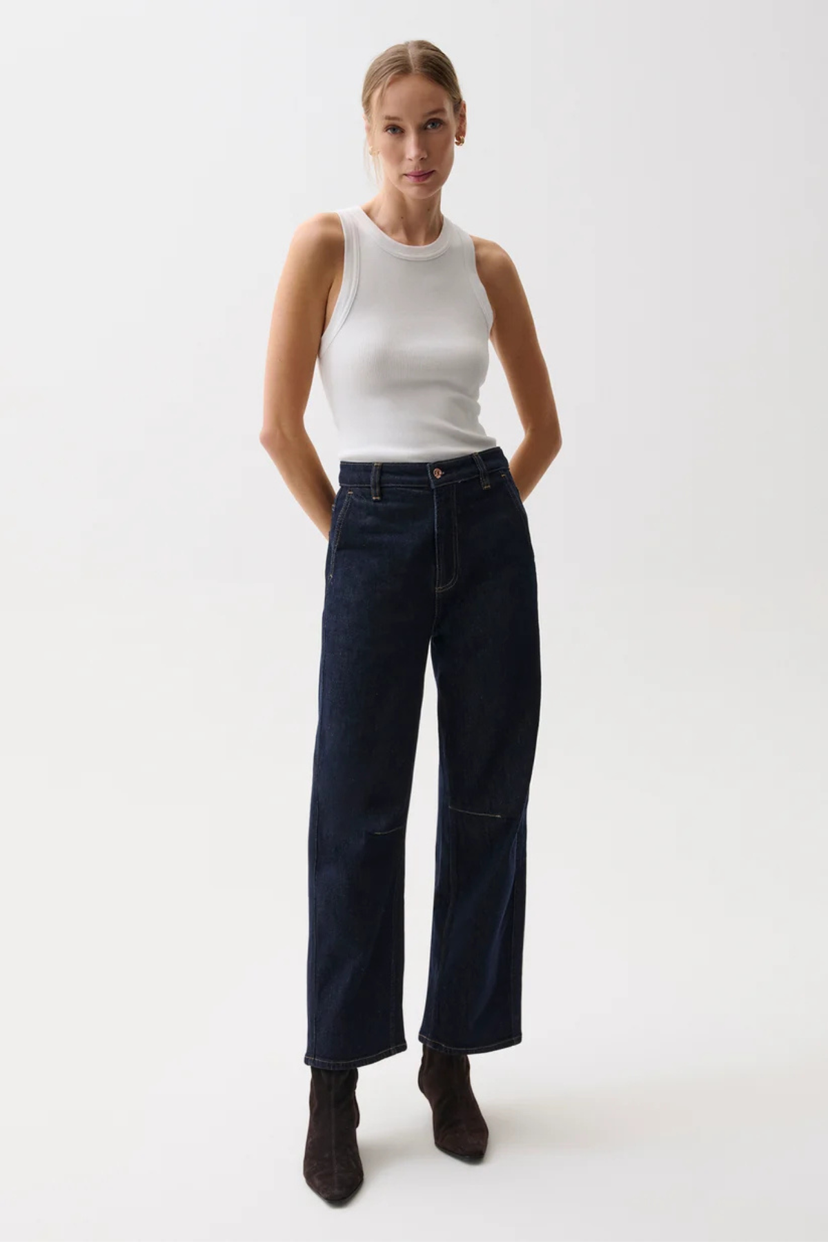 A woman modeling a pair of dark blue wide-leg denim jeans with a white sleeveless top and dark brown ankle boots, standing against a plain white studio background.