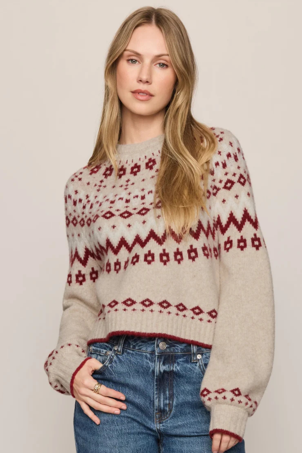 A woman modeling a cream knit sweater with a burgundy Fair Isle pattern, paired with blue jeans, against a solid light beige background.