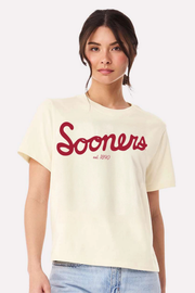 A woman wearing a cream-colored short-sleeve t-shirt with 'Sooners est. 1890' printed in red script across the chest, paired with light blue jeans, set against a plain light gray background.
