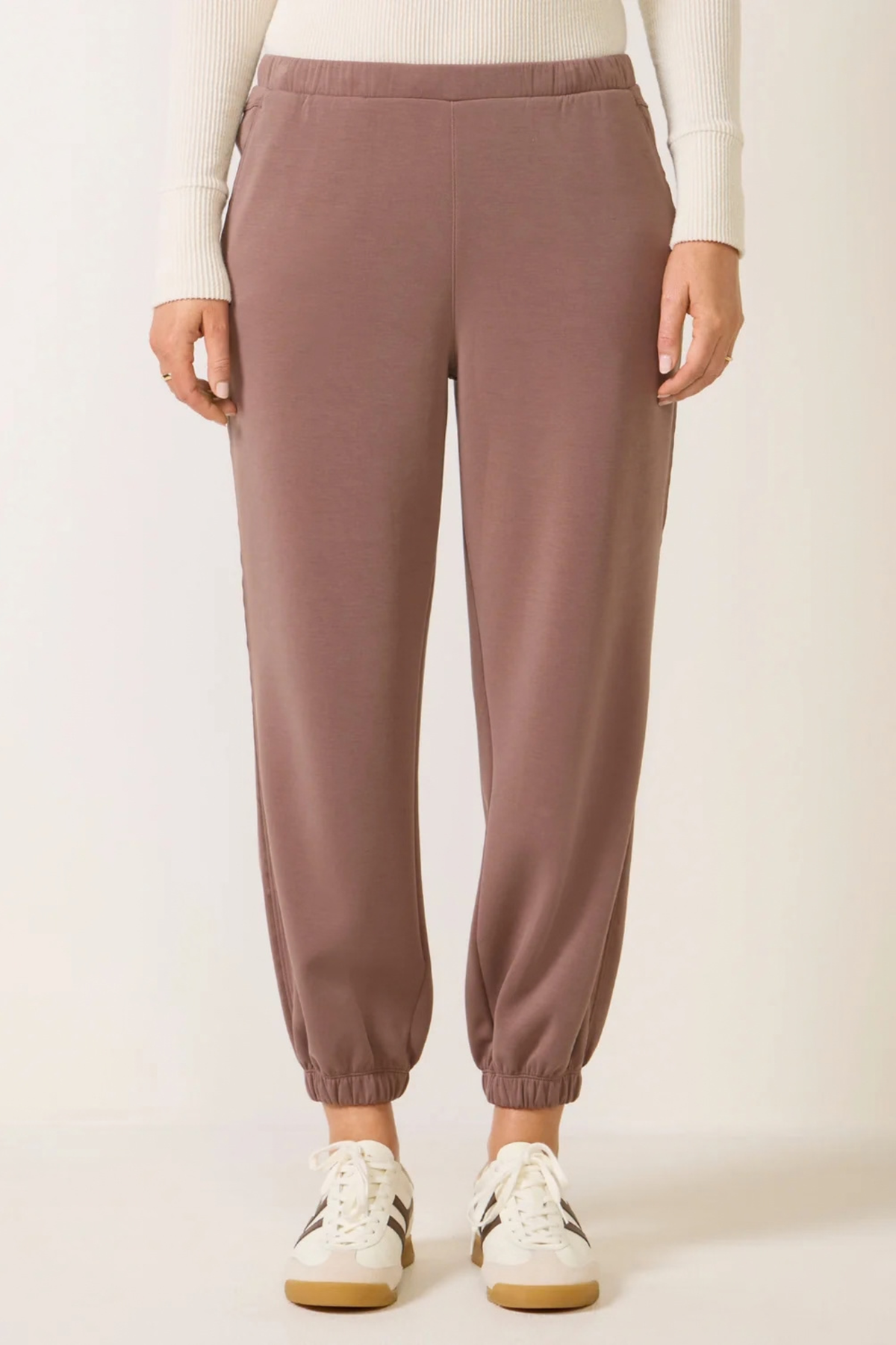 A person wearing dusty mauve jogger-style pants with an elastic waistband and elastic cuffs, paired with a white ribbed top and white sneakers, shown against a plain light background.