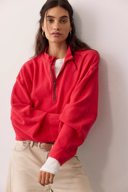 A woman wearing a bright red ribbed quarter-zip sweatshirt over a white undershirt and beige pants, leaning against a light gray wall.