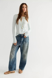A woman modeling a white long-sleeve crewneck top and wide-leg faded blue jeans with brown sandals, standing against a plain light gray studio background.