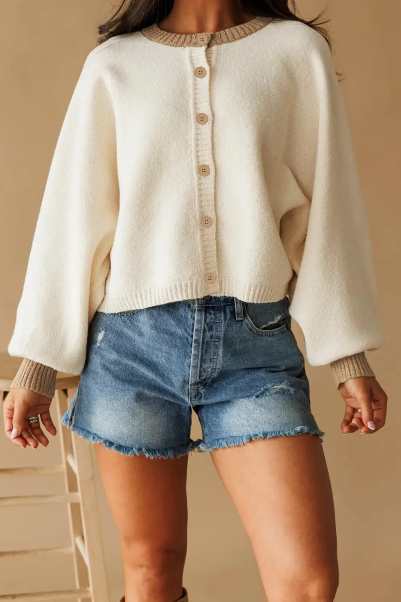 A woman wearing a cream-colored knit cardigan with beige ribbed trim and buttons, paired with distressed denim shorts, shown against a warm beige background.