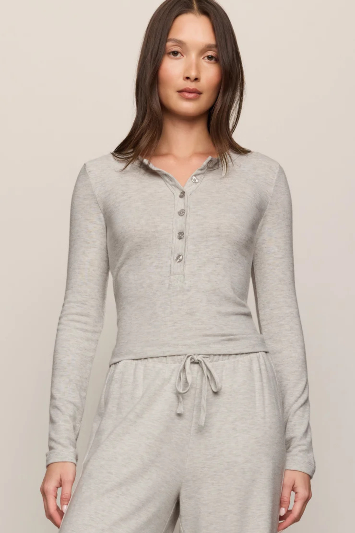 A woman modeling a light heather gray long-sleeve henley top with matching drawstring sweatpants against a plain beige studio background.