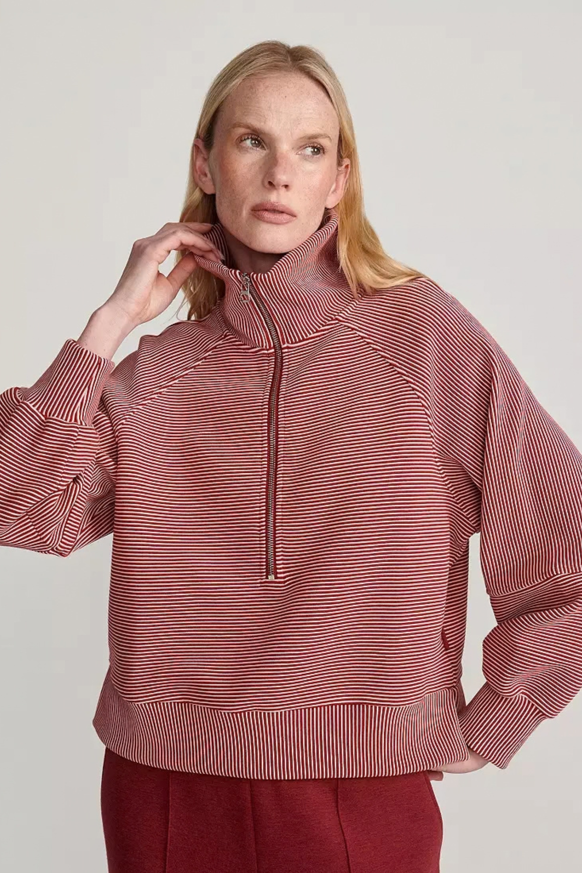 A woman wearing a cropped, long-sleeve quarter-zip pullover with fine horizontal stripes in dusty rose and white, paired with matching dark red pants, against a light gray studio background.