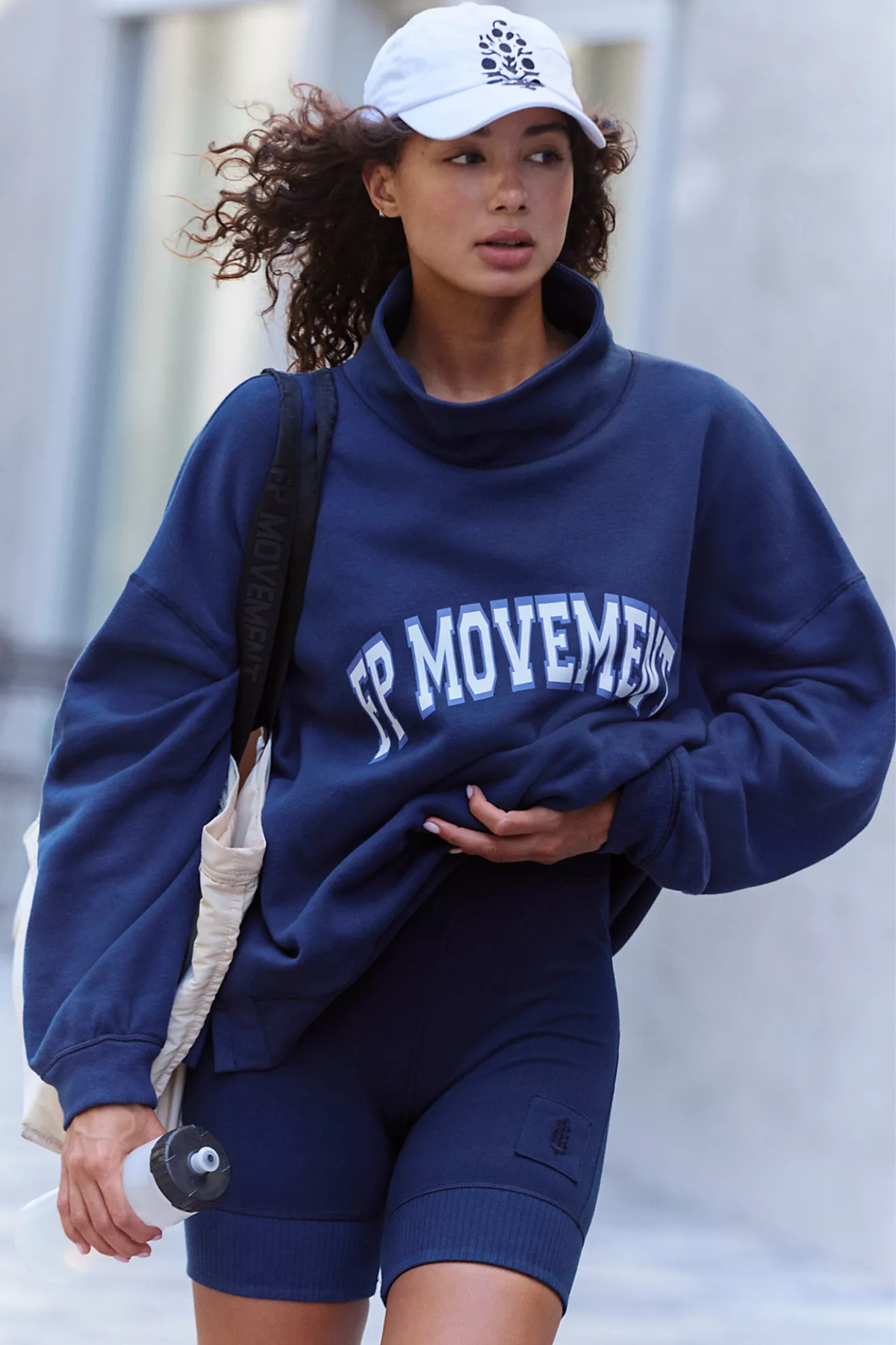 A woman wearing a navy blue turtleneck sweatshirt with 'JP MOVEMENTS' printed in white across the chest, matching navy biker shorts, and a white baseball cap, carrying a beige shoulder bag.