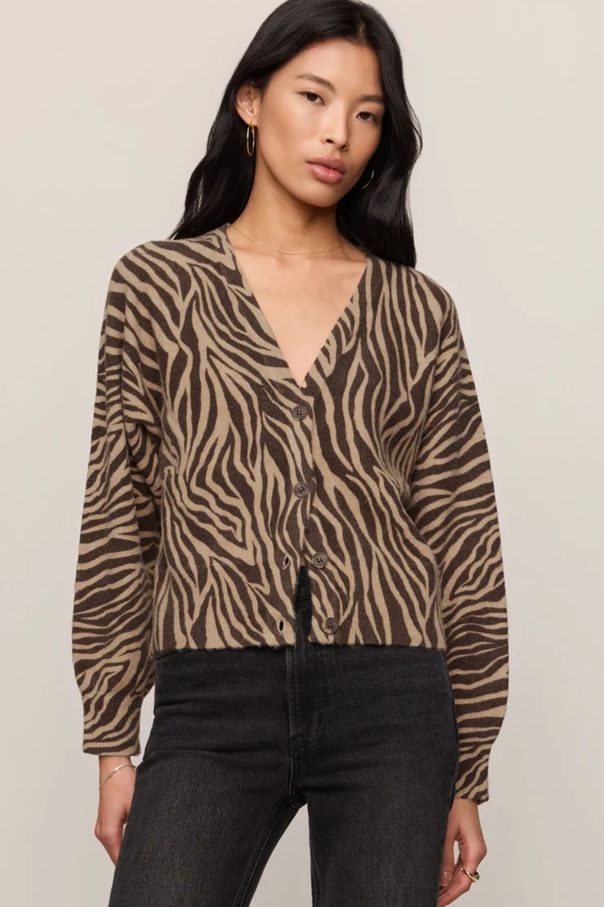 A woman modeling a brown and beige zebra-print cardigan with a V-neck and button-front closure, paired with black jeans, against a plain light beige studio background.