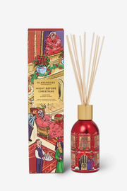 A reed diffuser bottle with natural reeds and a gold cap stands beside its illustrated retail box. The bottle is glossy red with a festive holiday illustration, and the box shows a warm Christmas interior scene with people and holiday decor.