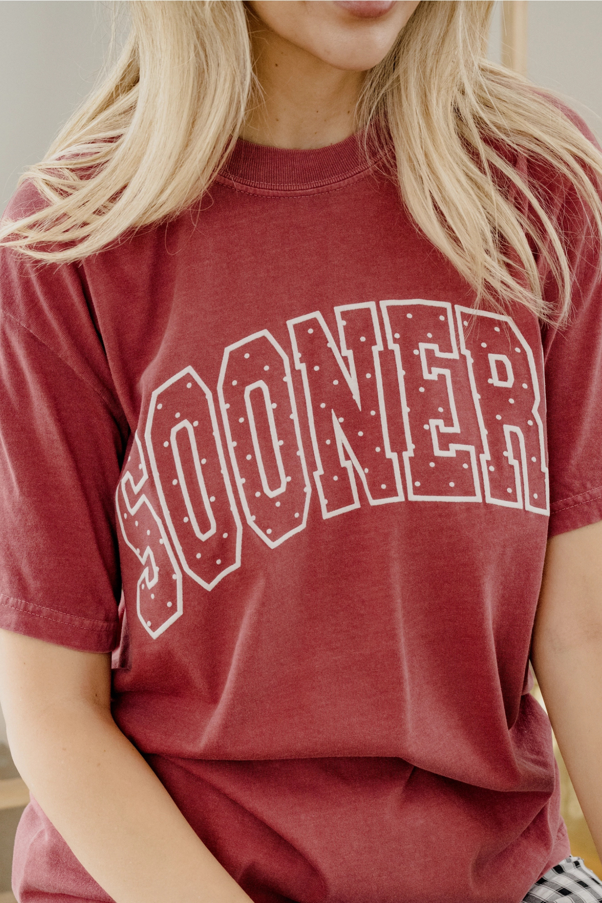 A close-up of a person wearing a faded red t-shirt with 'SOONER' printed in white arched block letters with small dot details inside the lettering.