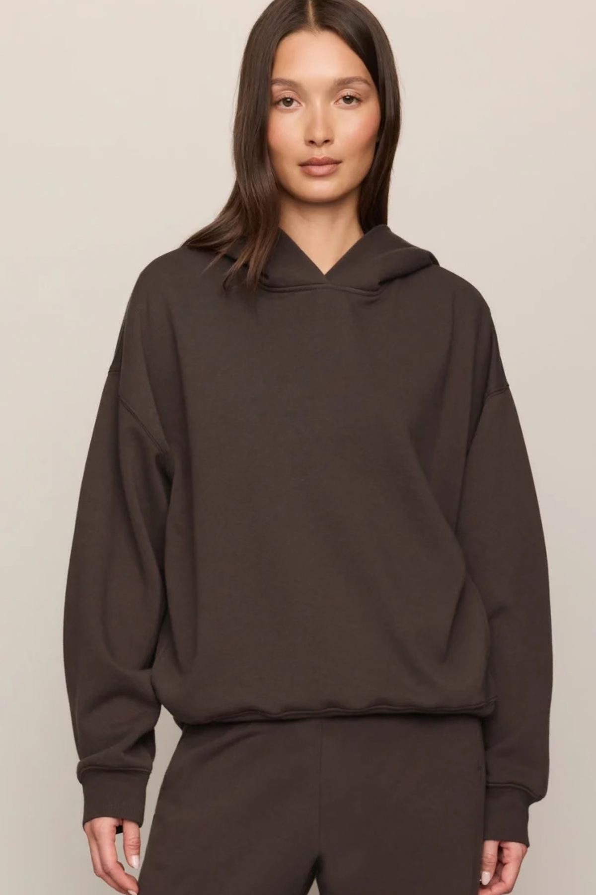 A woman wearing an oversized dark brown pullover hoodie and matching sweatpants, photographed against a plain light beige studio background.