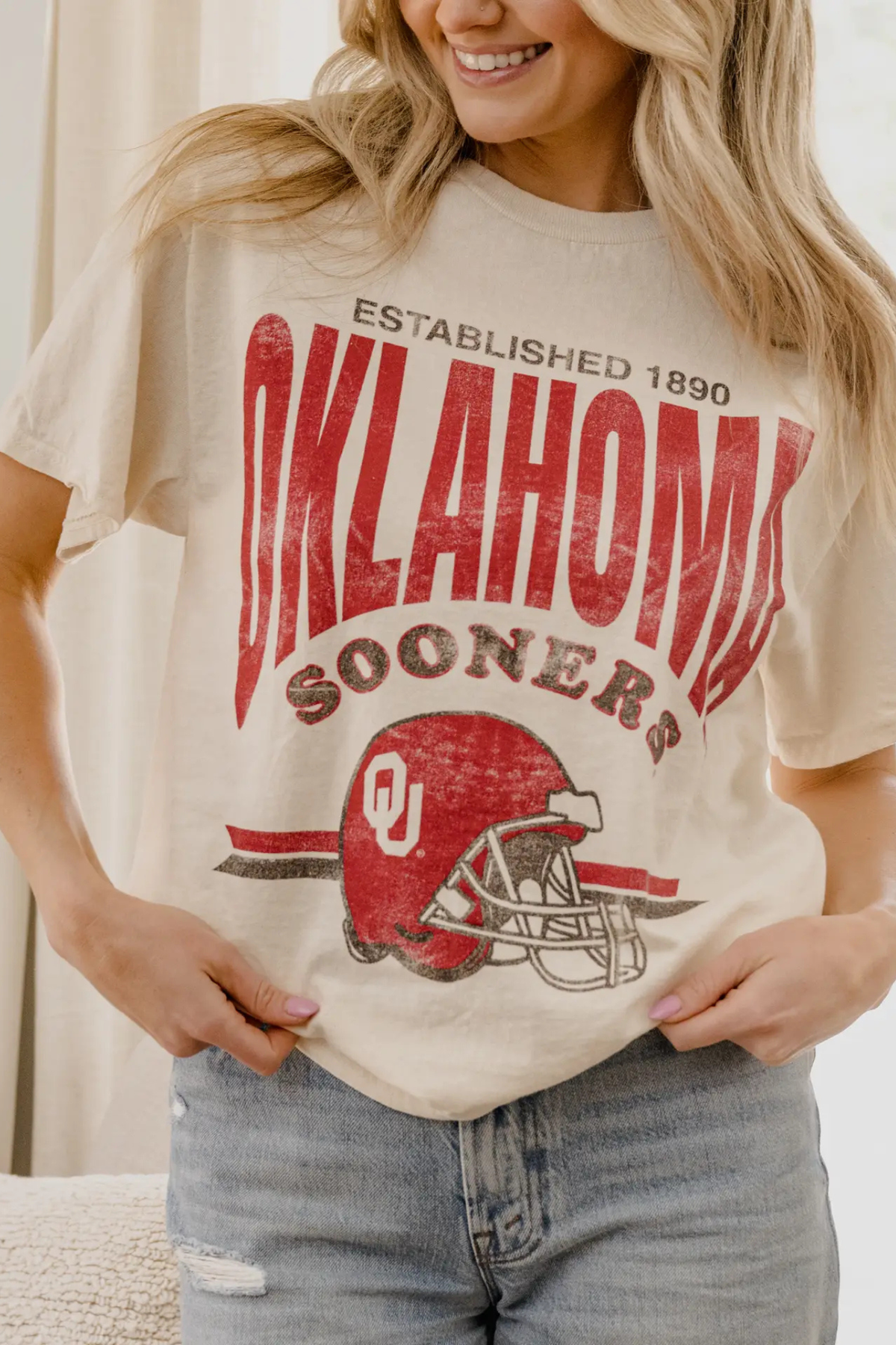 A woman wearing a cream-colored graphic t-shirt with a distressed red and brown print that reads 'OKLAHOMA SOONERS' and features an Oklahoma Sooners helmet logo. She is also wearing light-wash blue jeans.