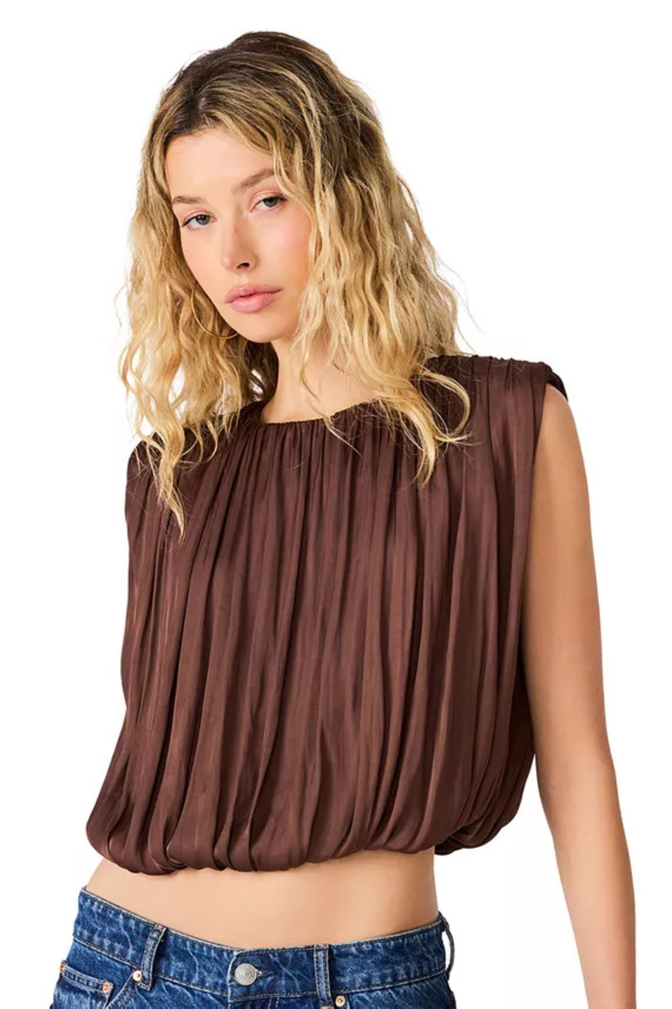 A woman with blonde wavy hair wearing a brown sleeveless top with a gathered, pleated texture and blue jeans, photographed against a solid white background.