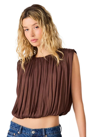 A woman with blonde wavy hair wearing a brown sleeveless top with a gathered, pleated texture and blue jeans, photographed against a solid white background.