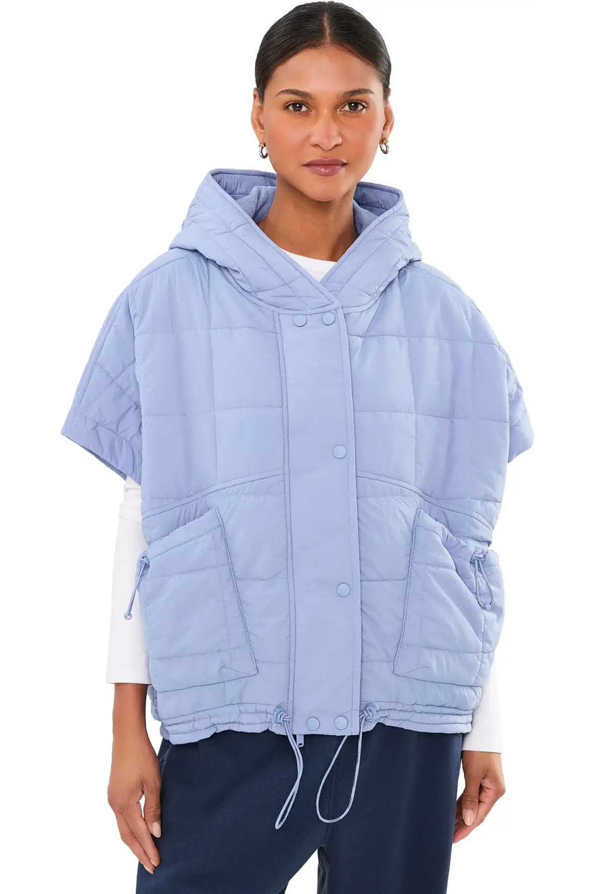 A model wearing a light blue hooded puffer vest with short sleeves, front snap buttons, and large front pockets, paired with a white long-sleeve shirt and navy pants, against a plain white background.