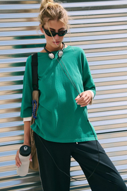 A woman wearing a vibrant green long-sleeve top and black pants, standing in front of a corrugated metal wall. She is carrying a shoulder bag and holding a water bottle.
