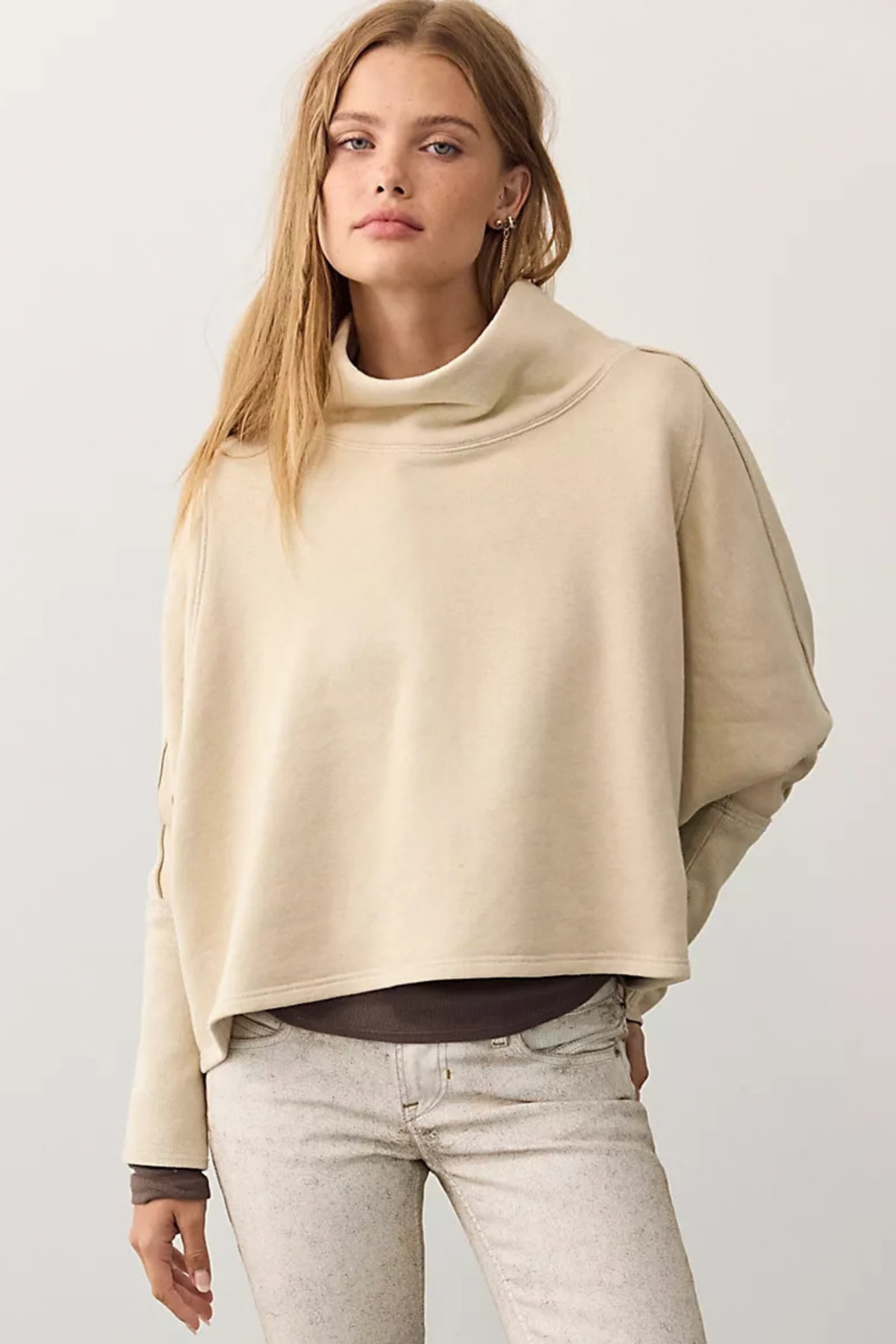 A woman wearing a beige cropped turtleneck sweatshirt with wide draped sleeves and light gray jeans, photographed against a plain light gray studio background.