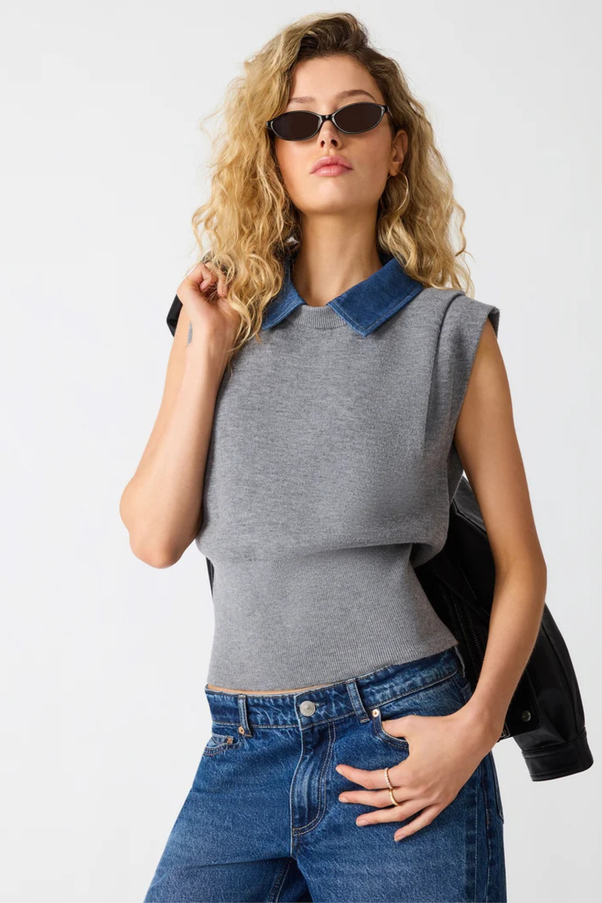 A woman modeling a gray sleeveless knit top with a blue denim collar, paired with blue jeans and black sunglasses, against a plain light gray background.