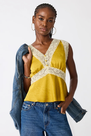 A woman modeling a sleeveless yellow top with cream lace trim and a buttoned V-neckline, paired with blue jeans and a draped denim jacket against a plain light background.