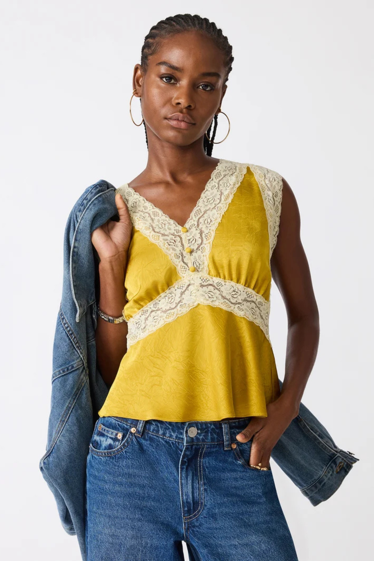 A woman modeling a sleeveless yellow top with cream lace trim and a buttoned V-neckline, paired with blue jeans and a draped denim jacket against a plain light background.