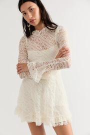 A woman modeling a short, long-sleeved lace dress in cream or off-white. The dress features a high ruffled collar, sheer patterned sleeves, and a ruffled hem, shown against a plain light gray studio background.