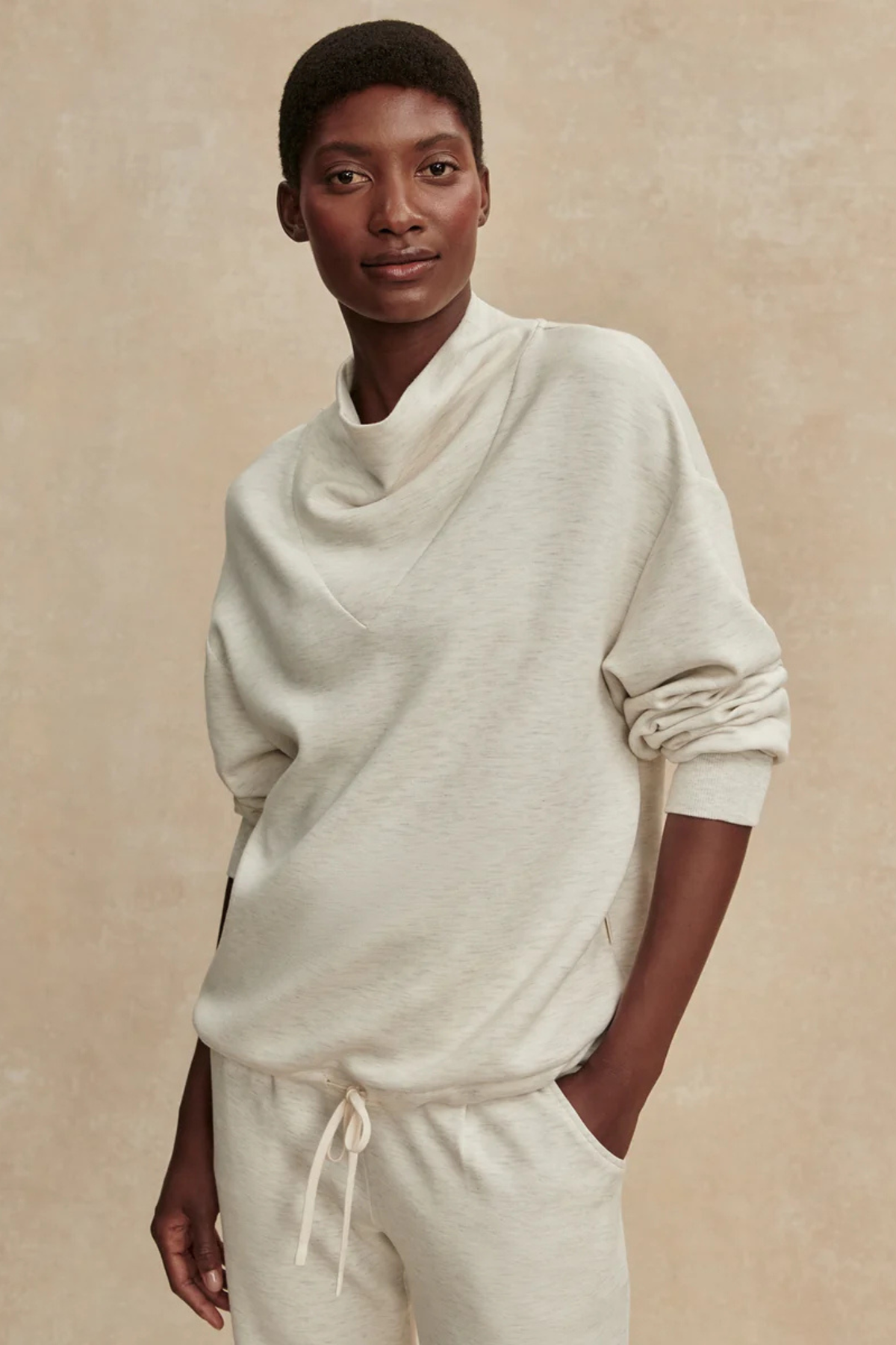 A model wearing a cream-colored cowl neck sweatshirt and matching sweatpants against a textured beige background.