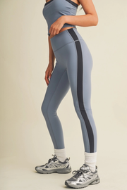 A model wearing high-waisted slate blue leggings with a bold black side stripe, paired with white socks and gray athletic sneakers, shown from the waist down against a solid beige background.