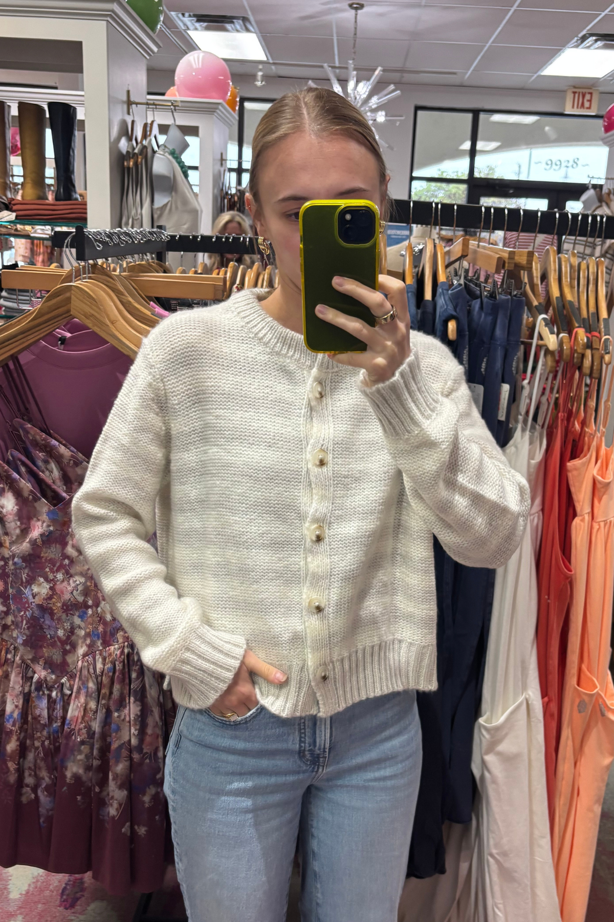 A woman takes a mirror selfie in a clothing store wearing a cream-colored, ribbed knit cardigan sweater with gold buttons and light blue jeans.